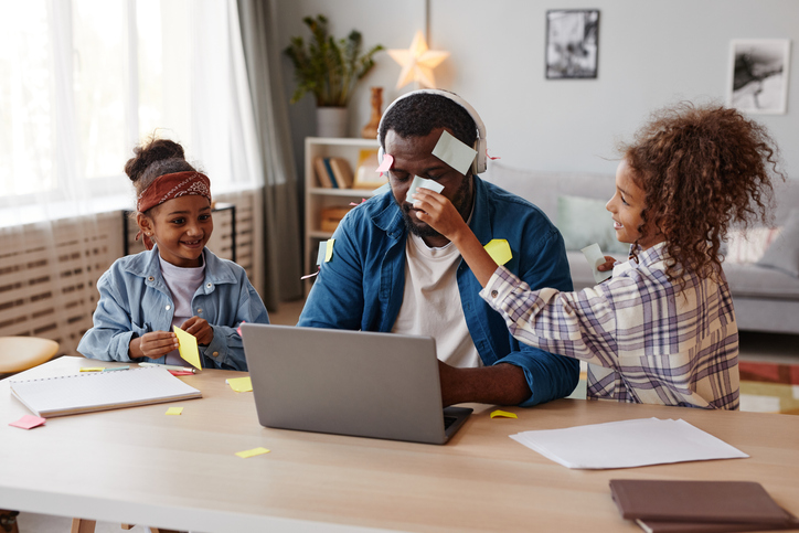 Father working from home on laptop and two children are putting sticky notes on him, disturbing his work